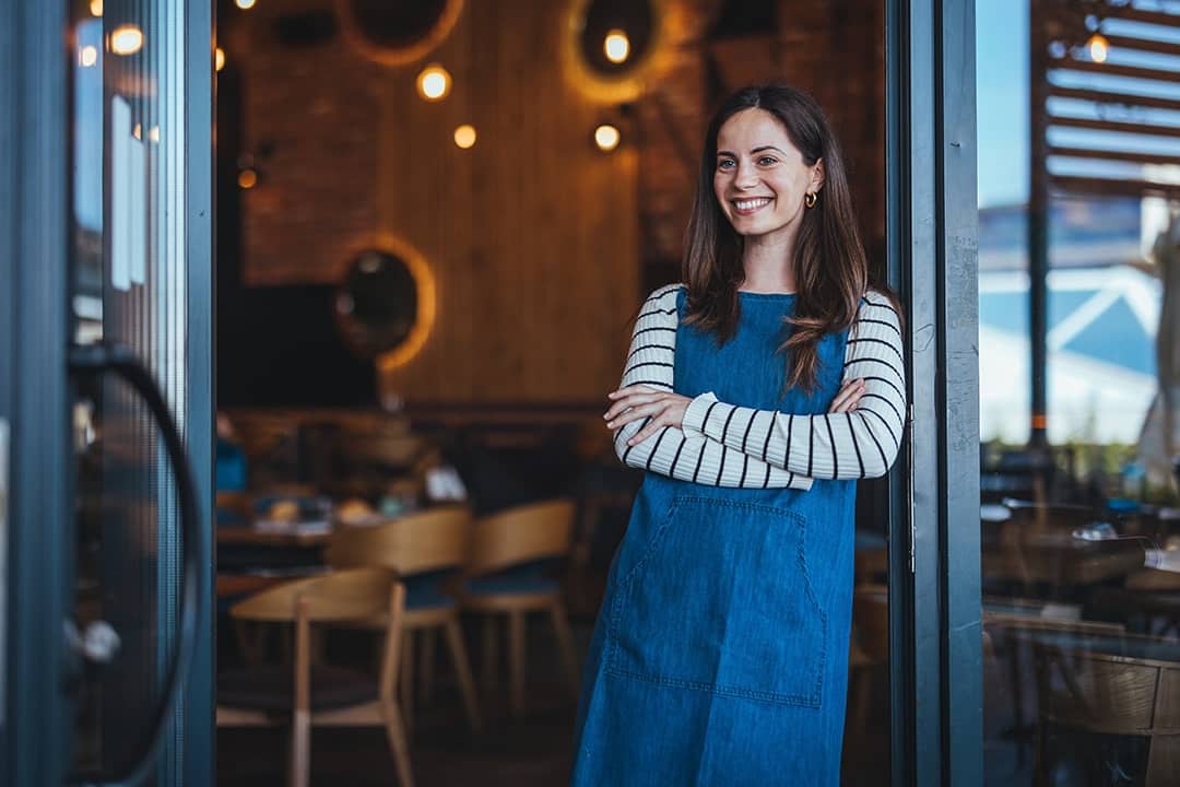 A female coffee shop owner happily stands at the entrance of her shop to welcome customers.